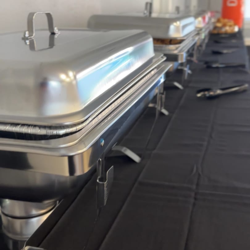 A close-up of a buffet line with several silver chafing dishes on a black tablecloth.