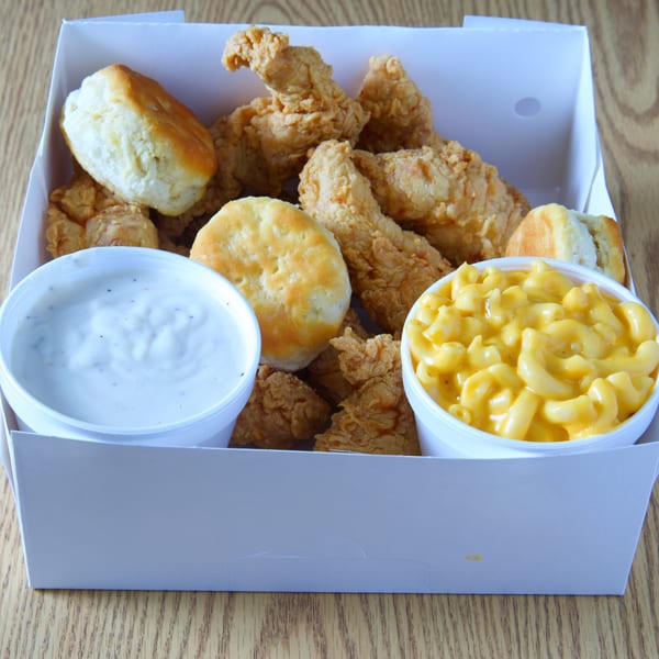 A white takeout box filled with fried chicken tenders, biscuits, mac and cheese, and dipping sauce.